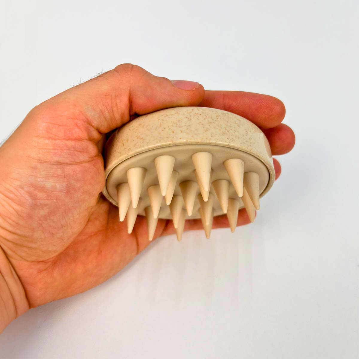  Ruka držiaca béžový silikónový masážny hrebeň na pokožku hlavy s mäkkými hrotmi, na bielom pozadí.